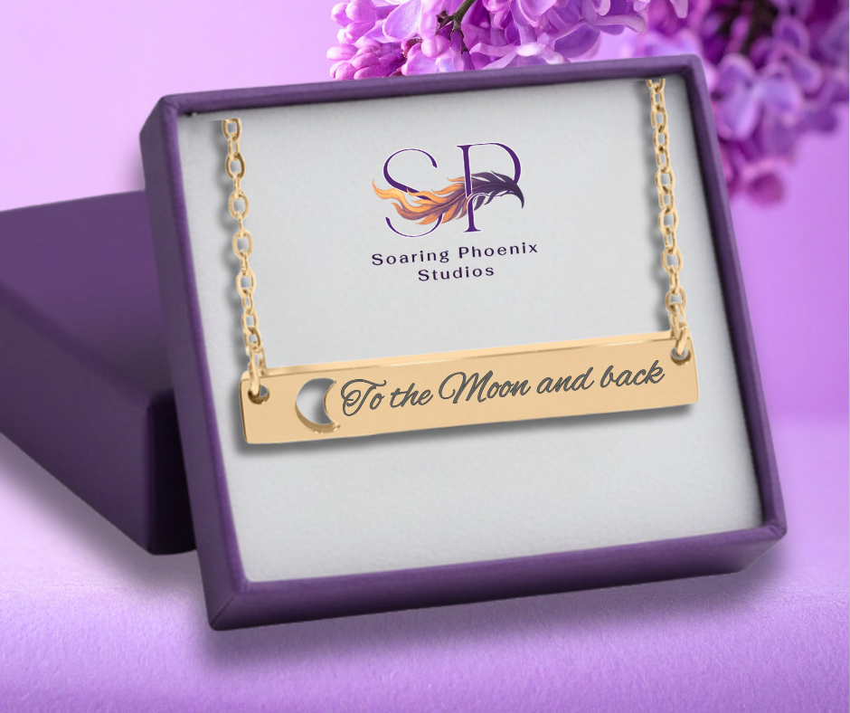 Gold necklace with 'To the Moon and back' engraving in a Soaring Phoenix Studios box on a purple background.